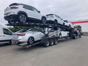 Car hauler loaded with white and silver SUVs and sedans on two decks, towed by a black Lee Bowe Company pickup truck.