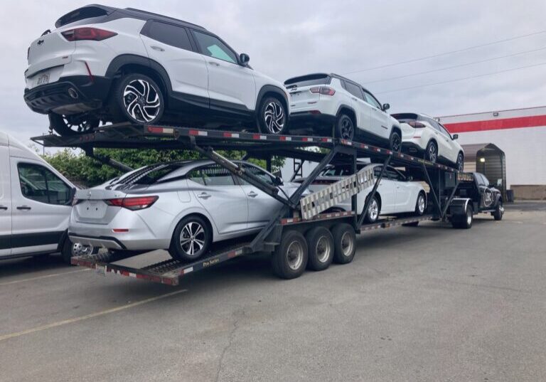 Car hauler loaded with white and silver SUVs and sedans on two decks, towed by a black Lee Bowe Company pickup truck.